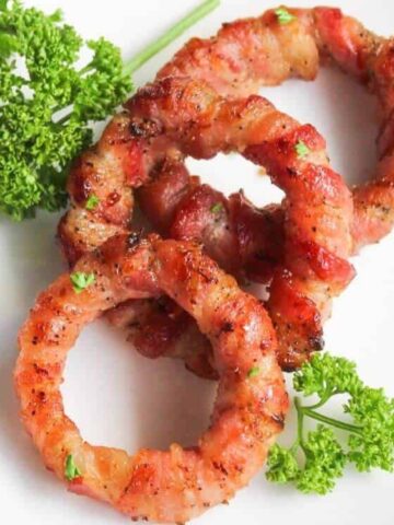 Three bacon-wrapped onion rings are arranged on a white plate, garnished with sprigs of fresh parsley.