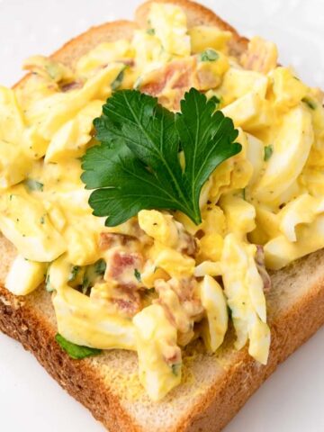 Slice of bread topped with egg salad and garnished with a sprig of parsley, served on a white plate.