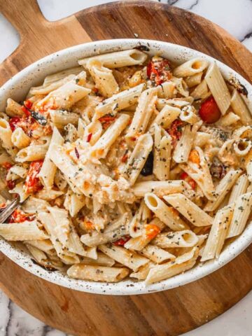 Oval dish filled with baked penne pasta, cherry tomatoes, and creamy sauce on a round wooden board with a fork; marble countertop and tomatoes visible.