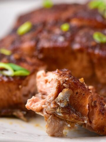 Close-up of a fork holding a piece of cooked, seasoned salmon fillet on a plate, garnished with sliced green onions.