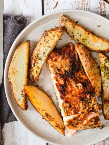 A plate with a serving of baked salmon and several seasoned potato wedges, next to a fork and knife on a gray napkin.