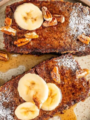 Two slices of French toast topped with banana slices, chopped pecans, powdered sugar, and syrup on a beige plate with two forks.