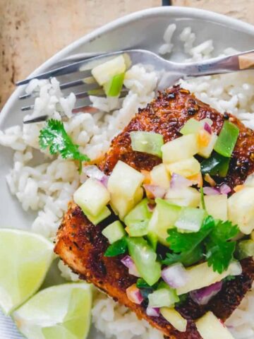 A plate of white rice topped with a grilled salmon fillet, pineapple salsa, and cilantro, served with lime wedges and a fork.