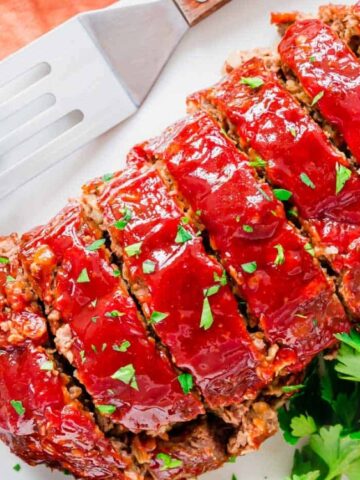 Sliced glazed meatloaf on a white board, garnished with fresh parsley, with a metal spatula and a cloth napkin nearby.