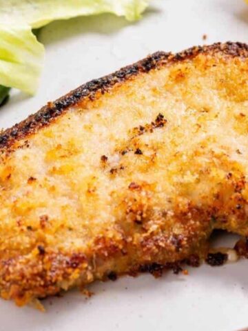 A breaded and baked fish fillet on a plate, served with green beans, lettuce, and a lemon wedge.