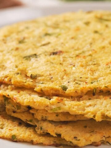 A close-up of several stacked, round, yellow corn tortillas with visible green herb flecks on a white plate.