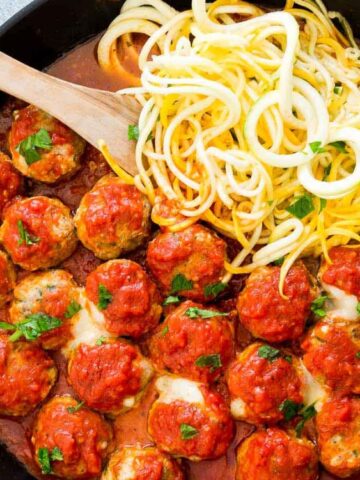 A skillet filled with meatballs in tomato sauce and zucchini noodles, garnished with chopped parsley, with a wooden spoon resting in the pan.