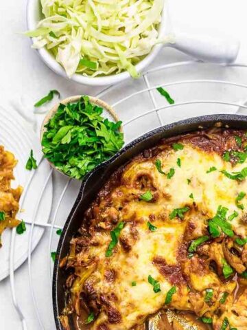A skillet of baked cheesy cabbage casserole sits beside a plate with a serving of the dish, a bowl of shredded cabbage, and chopped herbs on a white surface.