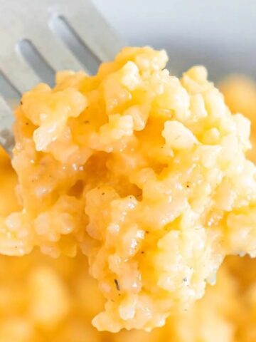 A close-up of a fork holding a bite of creamy, yellow macaroni and cheese, with more macaroni and cheese in the background.