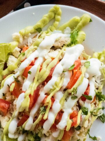 A plate of shredded chicken topped with diced tomatoes, chopped herbs, white and green sauces, and a lime wedge, served with a fork and knife on a wooden tray.