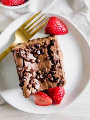A piece of chocolate chip cake with strawberries on a white plate, accompanied by a gold fork, on a light napkin.