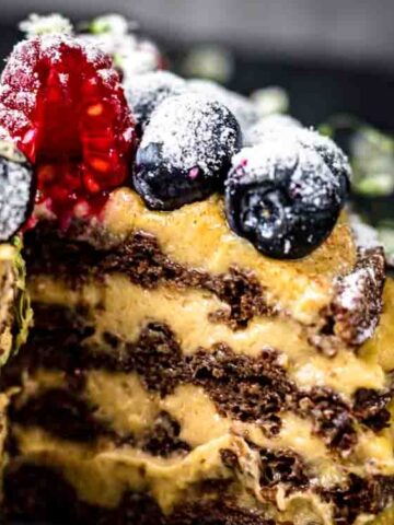 A layered chocolate and cream dessert topped with blueberries, raspberries, and powdered sugar, with a slice removed to show the interior.