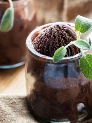 Two glass jars filled with chocolate ice cream, each garnished with fresh green leaves, placed on a brown cloth and wooden surface.