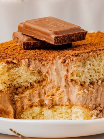 A slice of chocolate tiramisu topped with two pieces of chocolate sits on a white plate.