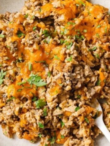 A bowl of ground beef and rice casserole topped with melted cheddar cheese and chopped herbs, with a spoon on the side.