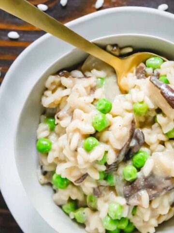 A bowl of risotto with green peas and mushrooms, served with a gold spoon on a wooden table, viewed from above.