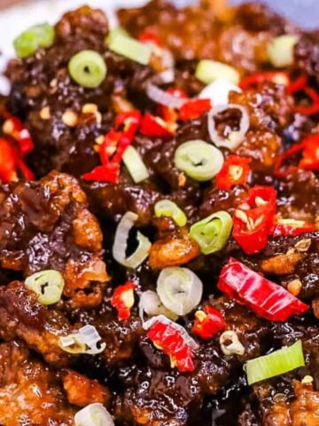 A plate of crispy fried beef coated in a dark sauce, garnished with chopped green onions and sliced red chili peppers.