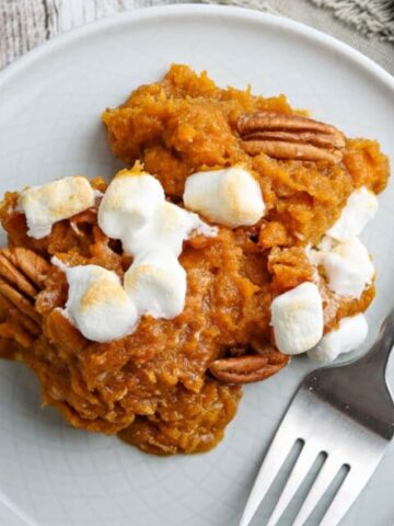 A serving of sweet potato casserole topped with pecans and mini marshmallows on a white plate with a fork.