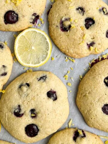 Large round cookies with visible blueberries are arranged on parchment paper. Lemon slices and grated lemon zest are also placed on the tray.
