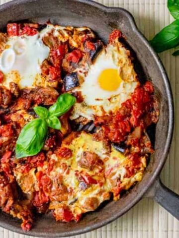A cast-iron skillet containing shakshuka with eggs poached in tomato and pepper sauce, garnished with fresh basil. A basil sprig is placed beside the skillet.
