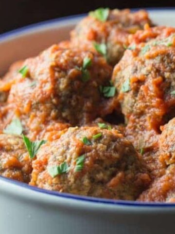 A white dish filled with meatballs covered in tomato sauce and garnished with chopped green herbs.