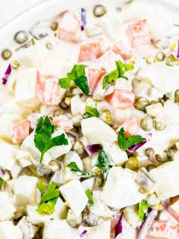 A plate of creamy potato salad with peas, carrots, red onion, and parsley garnish, served with a fork and knife.