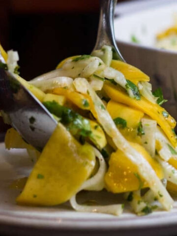 A pair of serving utensils places a portion of yellow and white vegetable salad with herbs onto a plate, with a bowl of more salad in the background.