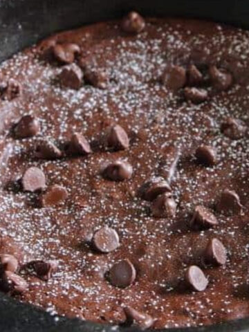 A chocolate chip skillet brownie is topped with powdered sugar and extra chocolate chips, shown in a black cast iron pan.