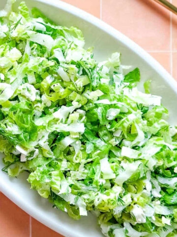 An oval white plate filled with finely chopped green lettuce sits on a peach-colored tiled surface next to two gold serving utensils.