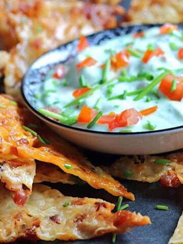 A bowl of creamy dip topped with chopped tomatoes and chives is surrounded by crispy, cheesy chips on a dark surface.