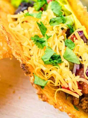 A crispy taco shell filled with seasoned ground beef, shredded cheese, lettuce, diced tomatoes, and chopped cilantro, with a side of fried fish and a bowl of diced red onions nearby.