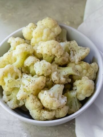 A white bowl filled with cooked cauliflower florets, seasoned with black pepper, sits on a light surface next to a white cloth.