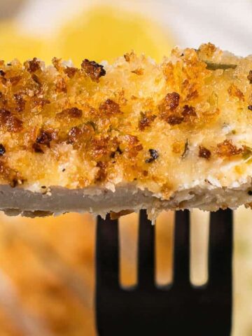 A fork holds a piece of breaded, baked fish fillet, showing a crispy golden-brown crust with a flaky white interior.