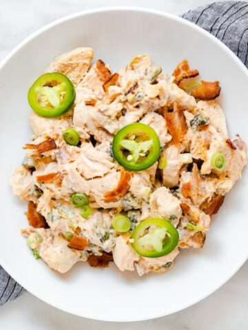 A white bowl filled with a creamy chicken dish, topped with sliced jalapeños, green onions, and bacon pieces, placed on a gray striped cloth.