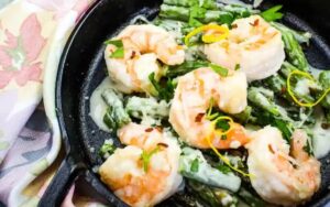 Shrimp and asparagus in a creamy sauce, garnished with parsley and lemon zest, served in a black skillet—perfect for those seeking delicious seafood recipes.