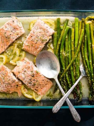 A glass baking dish with baked salmon fillets on lemon slices and roasted asparagus, held by two hands with a spoon resting in the dish.