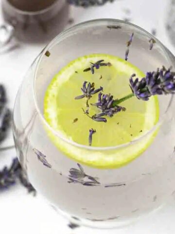 A glass of lemonade garnished with a lemon slice and a sprig of lavender, surrounded by dried lavender flowers on a white surface.
