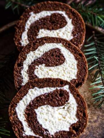 Three slices of chocolate and cream Swiss roll cake are arranged on a rustic plate, surrounded by green pine branches.