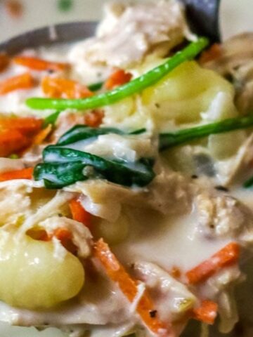 A close-up of a ladle holding creamy soup with gnocchi, shredded chicken, spinach, and carrots.