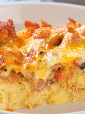 A close-up of a serving of breakfast casserole with melted cheese, bread, vegetables, and bits of meat on a white plate.