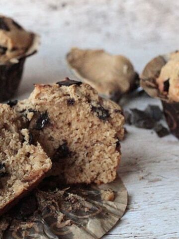Three chocolate chip muffins on a rustic surface, with one muffin cut in half to show its moist interior and chocolate chunks.