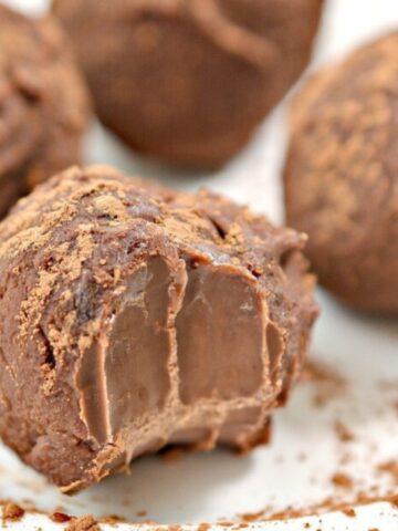 Four chocolate truffles sit on a white plate, one with a bite taken out to reveal a creamy interior. Cocoa powder is sprinkled on and around the truffles.