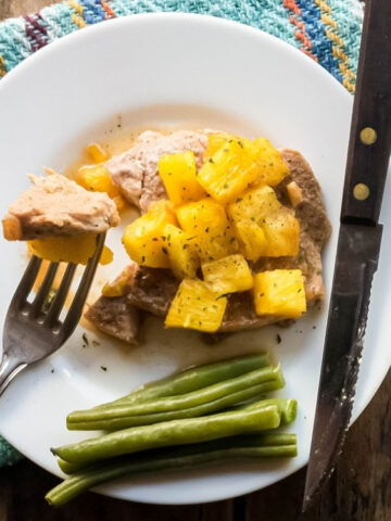 A plate with pork topped with pineapple chunks, green beans on the side, a knife, and a fork holding a piece of pork on a woven placemat.