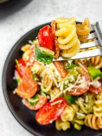 A fork holding a bite of pasta salad with tomatoes, green peppers, and cheese, above a black plate filled with the same salad.