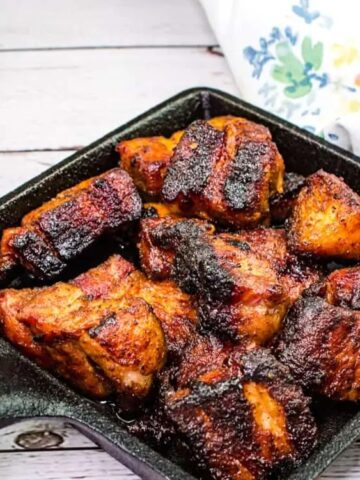 Grilled, seasoned chicken pieces in a cast iron skillet on a wooden surface, with a floral napkin and a yellow honey jar nearby.