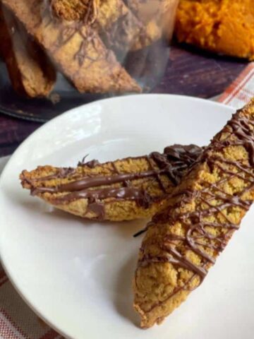 Two biscotti drizzled with chocolate sit on a white plate, with more biscotti in a glass jar and a spoonful of orange filling in the background.