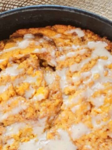 A baked cinnamon roll skillet cake with icing drizzled on top, shown in a black cast iron pan on a light surface with a piece of burlap and cinnamon sticks nearby.