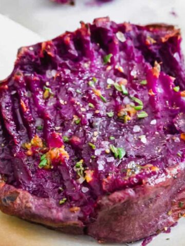 A baked smashed purple sweet potato topped with coarse salt and chopped herbs on parchment paper.