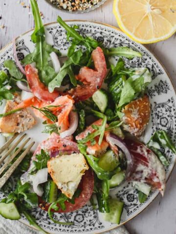 A plate with a partially eaten salad including greens, cucumber, red onion, tomato, cheese, and croutons, with a fork resting on the plate and a lemon half nearby.