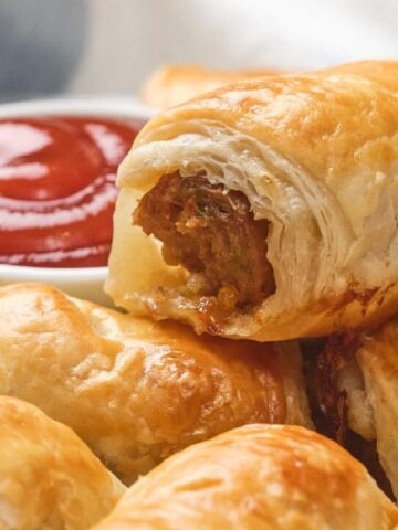 A close-up of golden puff pastry sausage rolls with a small bowl of tomato sauce in the background.